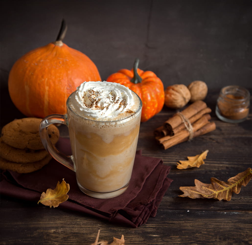 Latte de calabaza con crema batida y canela, acompañado de calabazas, ramas de canela y galletas en mesa otoñal.